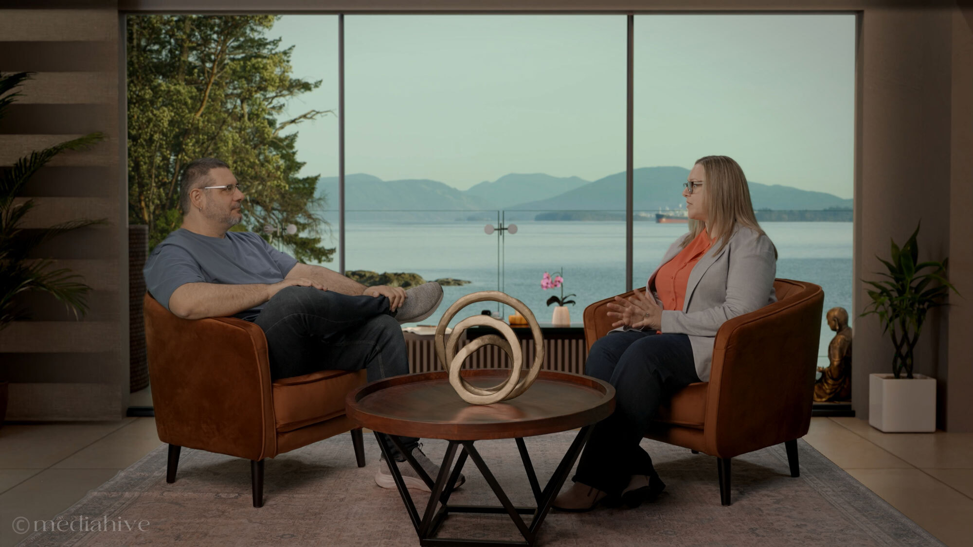 Two people seated in orange chairs on a MediaHive virtual set with a waterfront view